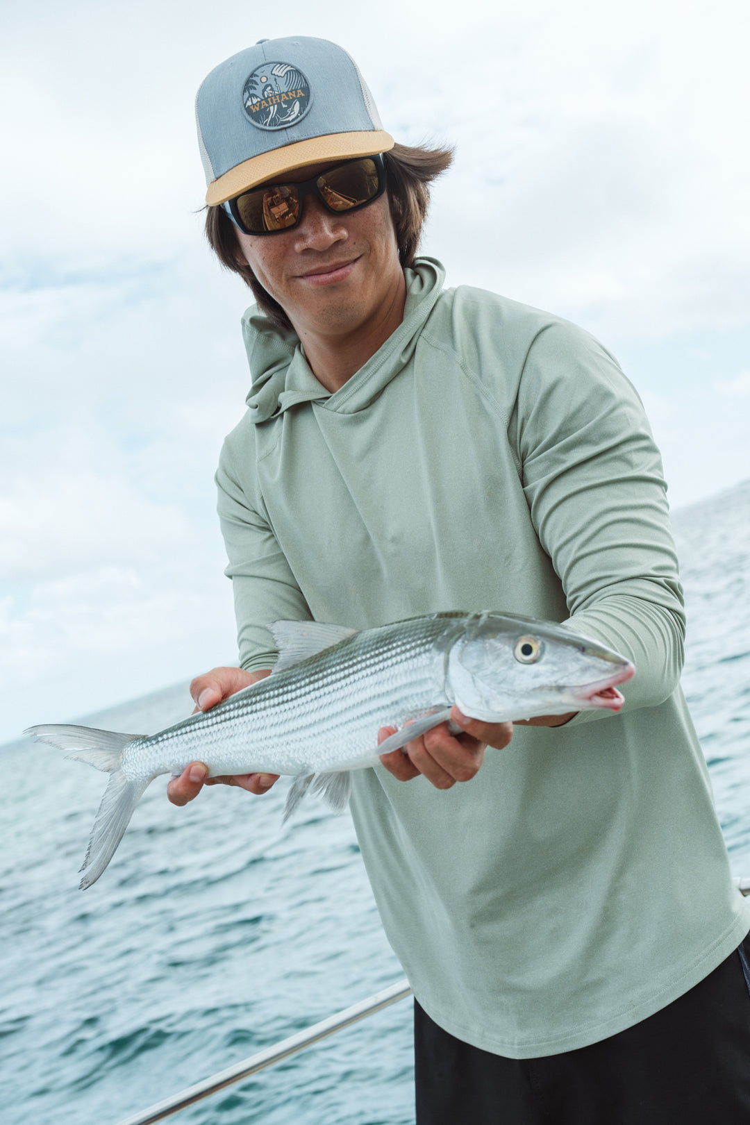 Smiling young man in a Waihana trucker hat and sage green hooded long-sleeve top holding a bonefish on a boat in Kaneohe Bay.